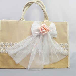 Könnte beinhalten: Eine beige Jute-Tragetasche mit weißem Spitzenbesatz und einer rosa Satinrose mit einer weißen Tüllschleife.