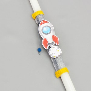 May include: A white candle decorated with a rocket ship, gray fabric, and yellow accents. The rocket is red, white, and blue with a cloud of smoke. A small blue flower charm hangs from the rocket.