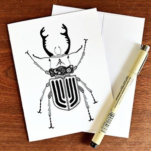 May include: A black and white line drawing of a beetle with intricate details on its body and large antlers. The beetle is drawn on a white card.