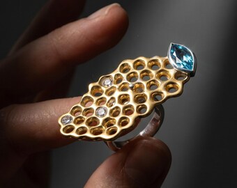 Puede incluir: Un anillo de tono dorado con una cara alargada de patrón de panal con recortes circulares. El diseño incluye tres pequeñas piedras redondas transparentes incrustadas en la red y una piedra preciosa azul de corte marquesa en el borde.