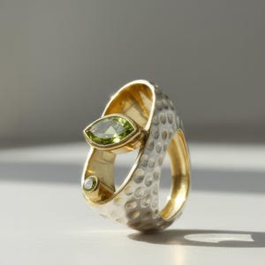 Puede incluir: Un anillo único con una gema verde talla marquesa engastada en oro. El anillo tiene una banda plateada texturizada con interior dorado y una gema redonda más pequeña. El diseño es moderno y artístico.