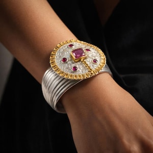 Può includere: Bracciale a polsino in argento con un grande elemento centrale ornato. L'elemento centrale presenta un bordo dorato, una gemma rosa centrale e gemme rosse più piccole. Il bracciale ha un design a coste.