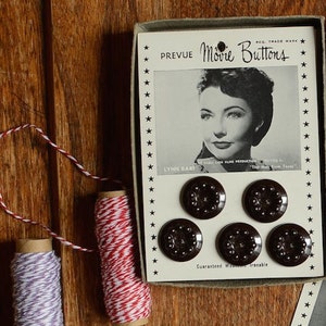 May include: A box of vintage "PREVUE Movie Buttons" featuring a black and white image of Lynn Bari. The box contains six dark brown buttons. Two spools of twine, one red and white, and one purple and white, are also visible.
