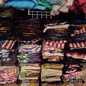 May include: A black wire shelf unit with multiple stacks of folded t-shirts. The t-shirts are in various colors and patterns, including red, white, blue, green, and black. Some of the t-shirts have graphic designs.