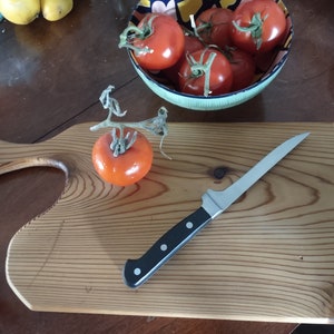 May include: A wooden cutting board with a handle, a knife, and a red tomato. The cutting board has a natural wood grain and is made of pine. The knife is a stainless steel boning knife with a black handle. The tomato is ripe and has a green stem.