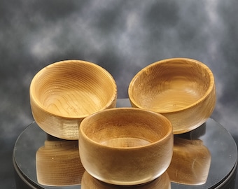 May include: A set of three small, round wooden bowls with a natural light brown finish. Each bowl features a smooth, polished surface that highlights the distinct, circular grain patterns of the wood. The bowls are arranged in a cluster.