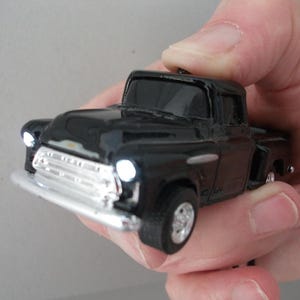 May include: A black toy pickup truck with a silver grill and headlights. The miniature vehicle is held in a hand, showcasing its glossy finish and detailed design. The truck's wheels and body are black, with silver accents.