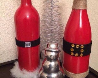 May include: Two red glass bottles decorated for Christmas. One bottle has a white fur trim and a black belt with a silver buckle. The other bottle has a brown twine wrap and a black belt with gold studs. A silver snowman ornament is in front of the bottles.