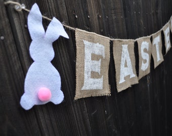 May include: A burlap banner with the word "Easter" in white letters, decorated with two white felt bunnies with pink pom-poms.