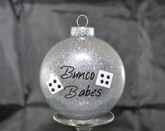 May include: Silver glitter Christmas ornament with black lettering that reads "Bunco Babes" and two black dice with white dots.
