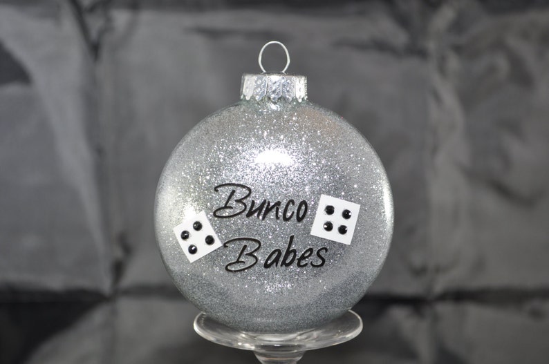 May include: Silver glitter Christmas ornament with black lettering that reads "Bunco Babes" and two black dice with white dots.