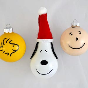 May include: Three Christmas ornaments featuring characters from the Peanuts comic strip. One ornament is yellow with a black outline of Snoopy's head. Another ornament is white with a black outline of Snoopy's face and a red Santa hat. The third ornament is beige with a black outline of Charlie Brown's face.