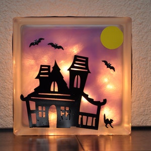 May include: A glass block with a light purple background and a silhouette of a haunted house, bats, and a yellow moon. The block is illuminated from behind, creating a glowing effect.