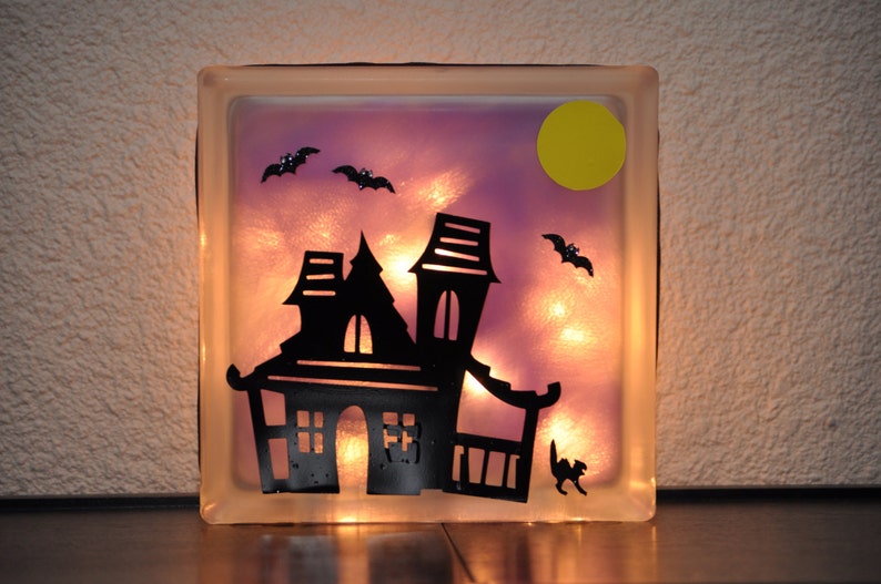 May include: A glass block with a light purple background and a silhouette of a haunted house, bats, and a yellow moon. The block is illuminated from behind, creating a glowing effect.
