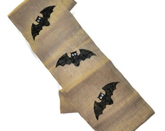 May include: A burlap table runner with three black bats with white eyes printed on it. The bats are flying in a horizontal position. The runner is a light brown color with a darker brown stripe running down the center.