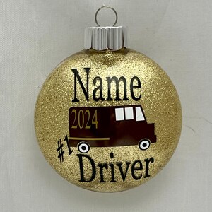 May include: Gold glitter Christmas ornament with a brown school bus, the number 2024, and the text "#1 Driver" on it.