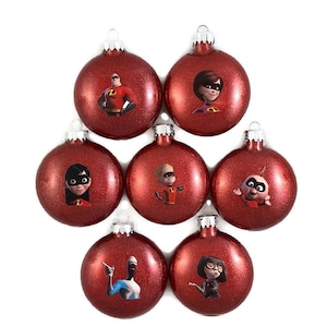 May include: Set of seven red glitter Christmas ornaments featuring characters from the animated film "The Incredibles". The ornaments feature Mr. Incredible, Elastigirl, Violet, Dash, Jack-Jack, Frozone, and Edna Mode.
