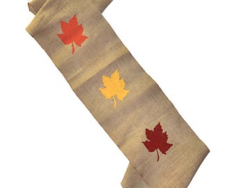 May include: A beige burlap table runner with three painted maple leaves. The leaves are red, orange, and yellow.