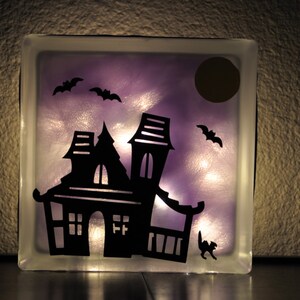 May include: A glass block with a purple background and a silhouette of a haunted house, bats, and a moon. The house has windows and a door. The block is lit from behind, creating a glow.