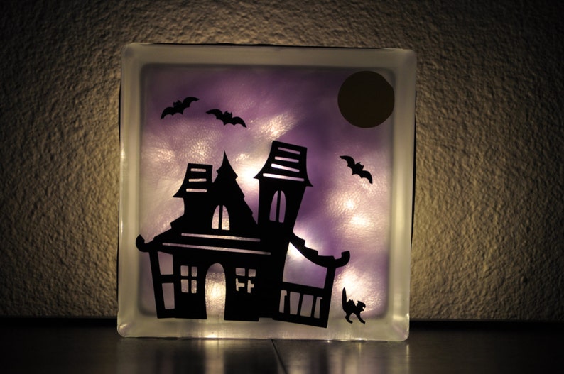 May include: A glass block with a purple background and a silhouette of a haunted house, bats, and a moon. The house has windows and a door. The block is lit from behind, creating a glow.