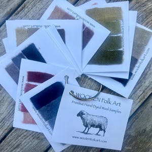 Puede incluir: Un abanico de muestras de tela de lana te&ntilde;ida a mano unidas a un anillo de metal. Cada tarjeta blanca presenta una tira de lana de colores en varios tonos incluyendo verde, azul, rojo y marr&oacute;n, con el texto Woolen Folk Art y una oveja.