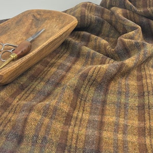 May include: A length of woven fabric featuring a plaid pattern in shades of brown, tan, and muted green. A wooden bowl with a rough, carved texture sits on the fabric, containing a pair of metal scissors with a wooden handle and a metal point.