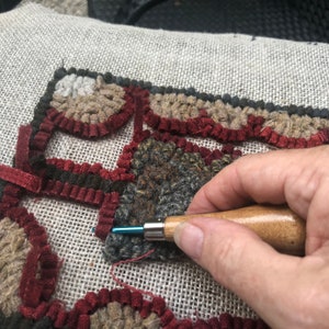 May include: Close-up of rug hooking in progress. The rug design features a geometric pattern with shades of brown, gray, and burgundy. A hand holds a wooden-handled hook, inserting it into the fabric to create the rug's texture.