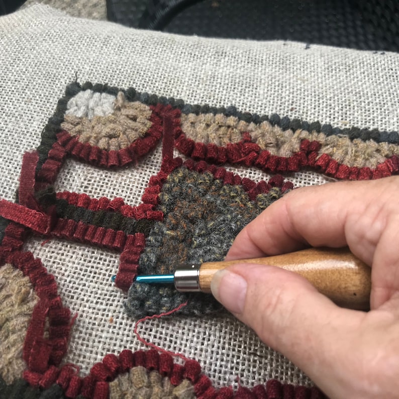 May include: Close-up of rug hooking in progress. The rug design features a geometric pattern with shades of brown, gray, and burgundy. A hand holds a wooden-handled hook, inserting it into the fabric to create the rug's texture.
