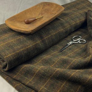 May include: A folded length of brown and green plaid fabric with yellow stripes. A wooden bowl with a small wooden and brass tool sits on top. A pair of silver scissors rests on the fabric.