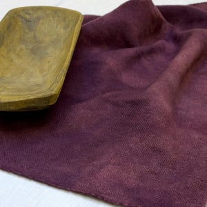 Puede incluir: Un camino de mesa de tela color ciruela intenso con una textura tejida visible y un patrón de tinte irregular y moteado, colocado junto a un cuenco de madera alargado y desgastado con un interior liso y cóncavo.