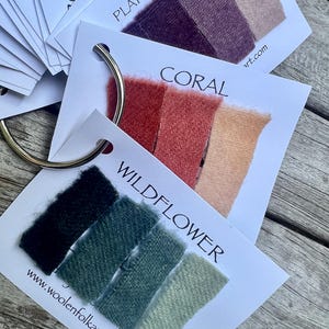 Puede incluir: Un conjunto de tarjetas de muestras de tela unidas por una anilla met&aacute;lica. Cada tarjeta presenta tres muestras rectangulares en tonos degradados, etiquetadas con nombres como WILDFLOWER, CORAL y PLATTER OF PLUMS, incluyendo woolenfolkart.com.