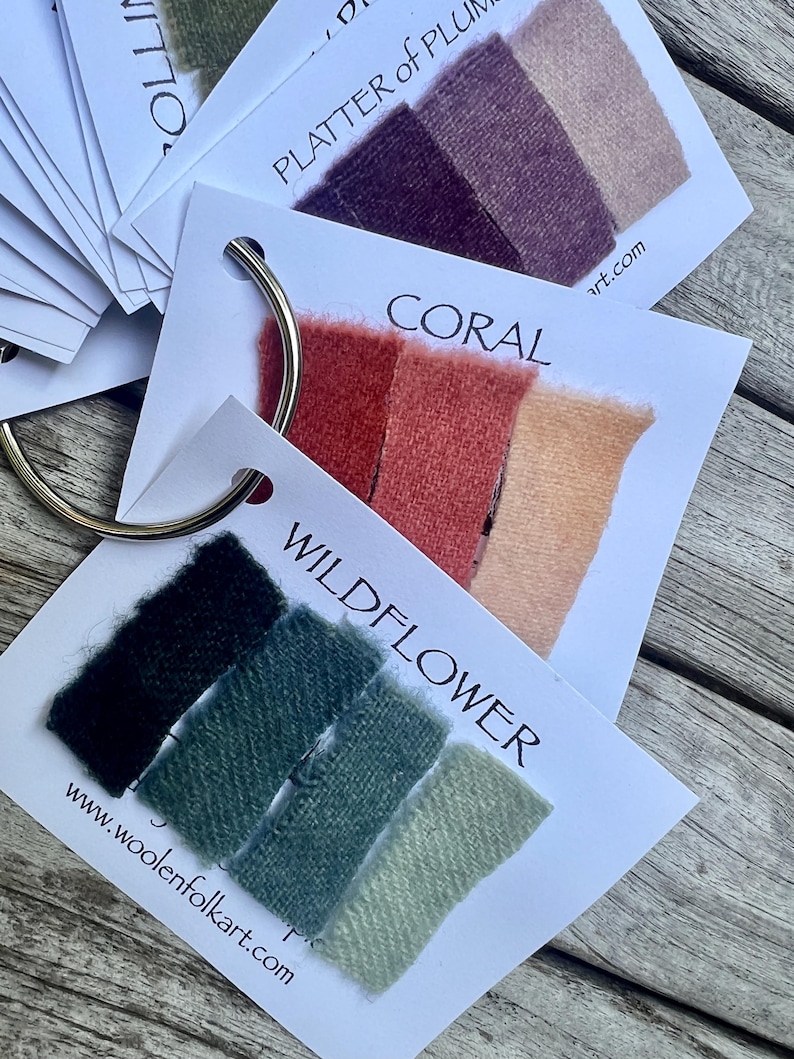 Puede incluir: Un conjunto de tarjetas de muestras de tela unidas por una anilla met&aacute;lica. Cada tarjeta presenta tres muestras rectangulares en tonos degradados, etiquetadas con nombres como WILDFLOWER, CORAL y PLATTER OF PLUMS, incluyendo woolenfolkart.com.