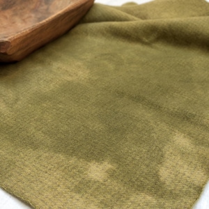 Puede incluir: Una pieza de tela verde oliva con un patrón de tinte moteado e irregular que presenta parches más claros por todo el tejido. Un pequeño cuenco de madera tallada de color marrón oscuro con una textura rugosa y natural se sitúa arriba a la izquierda.