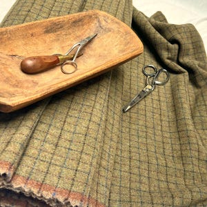 May include: A folded length of olive green wool fabric featuring a dark blue grid pattern, accompanied by a rustic wooden dough bowl and two pairs of silver metal sewing scissors with one pair having a wooden handle for craft projects.