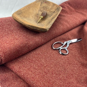 Puede incluir: Tela de espiga roja con una textura sutil. La tela está doblada y colocada sobre una superficie blanca. También se puede ver un cuenco de madera y unas tijeras en la imagen.