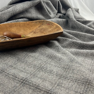 May include: A wooden dough bowl with a small awl and scissors rests on a gray woven fabric. The bowl has a natural wood grain and a curved shape. The fabric has a subtle herringbone pattern.