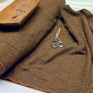 May include: A bolt of brown fabric featuring a woven herringbone pattern. A pair of silver metal scissors rests on the fabric, and a second pair of silver scissors is placed inside a small, shallow, oval-shaped wooden bowl.