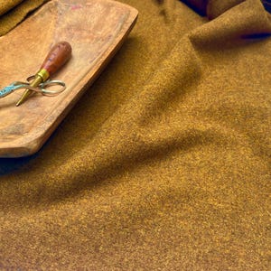 Puede incluir: Una tela texturizada de color marrón mostaza con una apariencia suave y afieltrada drapeada en pliegues, acompañada por una bandeja de madera que contiene unas tijeras de metal con mango marrón y una herramienta de metal puntiaguda con mango marrón a juego.