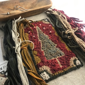 May include: A handcrafted rug with a Christmas tree design in red, green, and tan hues. The rug is surrounded by bundles of colorful fabric strips, a wooden bowl, and a pair of scissors. The rug measures approximately 10 inches by 8 inches.