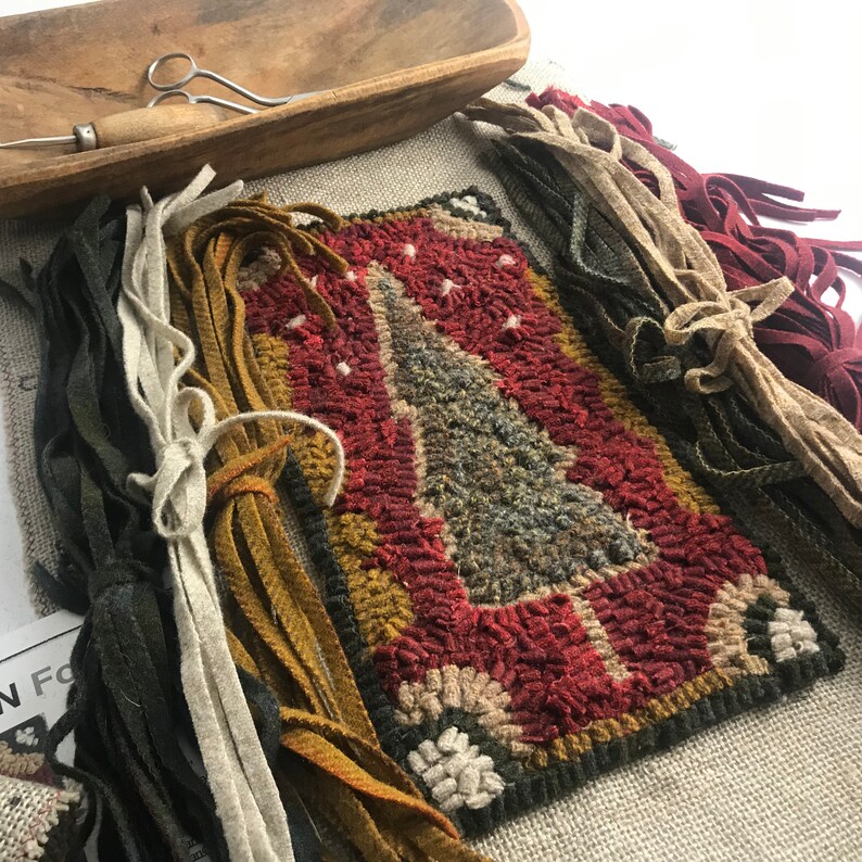 May include: A handcrafted rug with a Christmas tree design in red, green, and tan hues. The rug is surrounded by bundles of colorful fabric strips, a wooden bowl, and a pair of scissors. The rug measures approximately 10 inches by 8 inches.