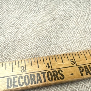 May include: Close-up of a light gray fabric with a diamond pattern. A wooden ruler with black text that reads "DECORATORS - PAINTED" is in the foreground, measuring up to 6 inches.