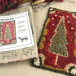 May include: A completed wool rug hooking project depicting a pine tree with a red, green, and brown color scheme. The rug is framed with a gold and white border. The image also includes a pattern for the rug, which is titled "WOOLEN Folk Art" and includes instructions for rug hooking or punch needle. The pattern is for a 6-inch by 12-inch strip of 1/4-inch primitive linen.
