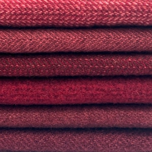 Puede incluir: Primer plano de seis muestras de tela en tonos rojos. Las texturas varían, incluyendo espiga y tejidos lisos. Las telas están apiladas, mostrando las diferentes texturas y tonalidades del material. La imagen destaca la variedad de la tela.