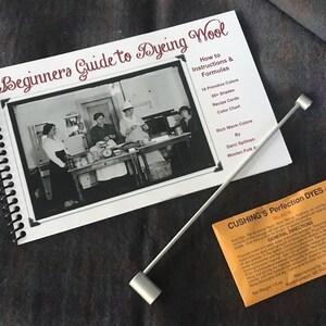 Puede incluir: Un libro de espiral titulado Beginners Guide to Dyeing Wool con una foto hist&oacute;rica en blanco y negro en la portada, una varilla mezcladora de metal plateado con extremos cil&iacute;ndricos y un peque&ntilde;o paquete naranja de tinte para lana.