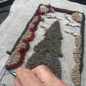 May include: A close-up of a rug hooking project in progress. The design features a stylized tree and border in shades of brown, red, and gray. The rug is being hooked on a burlap-like fabric with a blue and silver tool.