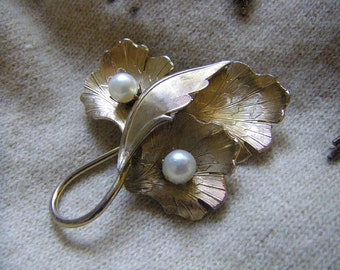 Peut inclure: Une broche en métal doré en forme de trois feuilles de ginkgo aux surfaces texturées en éventail. Deux perles blanches sphériques sont serties au centre de deux des feuilles. La broche présente une tige incurvée qui forme une boucle.