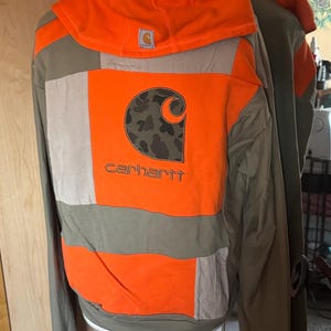 Puede incluir: Una sudadera con capucha naranja y verde oliva con un diseño de patchwork. La parte trasera presenta un gran logotipo de Carhartt con un patrón de camuflaje. Las mangas y la capucha son naranjas, y el cuerpo es verde oliva y beige.