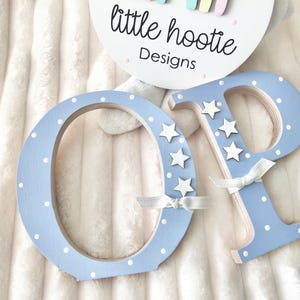 May include: Two wooden letters O and P painted light blue with white polka dots. Each letter features three small white star cutouts and a thin white ribbon bow. A circular white sign above displays the text little hootie Designs.