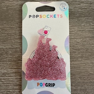 May include: A pink glitter PopSockets PopGrip in the shape of a castle, attached to a white and iridescent backing card. The card features the PopSockets logo and a graphic of a phone with a hand.