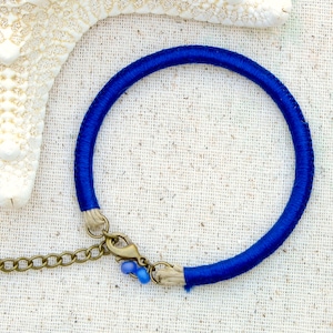 May include: A blue cord bracelet with a silver clasp and a chain attached. The bracelet is on a white fabric background.